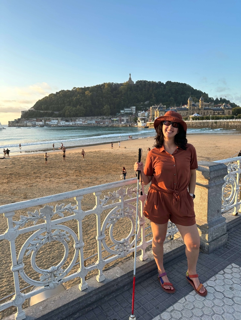 A woman holding a white and red cane stands in front of a white railing in San Sebastián, with the ocean and a hill in the background