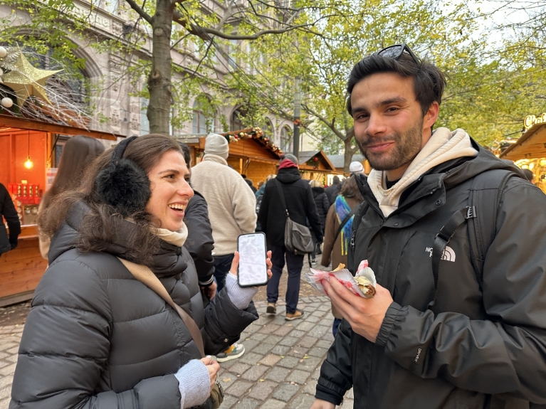 Two people laugh at a Christmas market, surrounded by trees and market booths. One of them is holding a crepe.