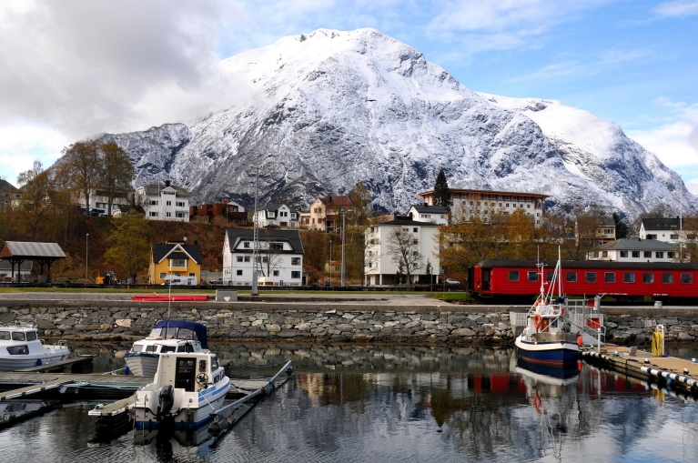 A red train alongside a small harbor with snowy mountains in the distance